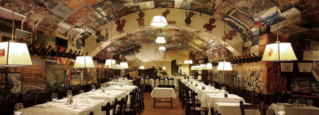 Interior de um restaurante com teto abobadado decorado com pôsteres e desenhos de crianças. Mesas retangulares com toalhas brancas e cadeiras de madeira escuras estão alinhadas em duas fileiras, com talheres e taças. Luminárias pendentes iluminam o ambiente aconchegante