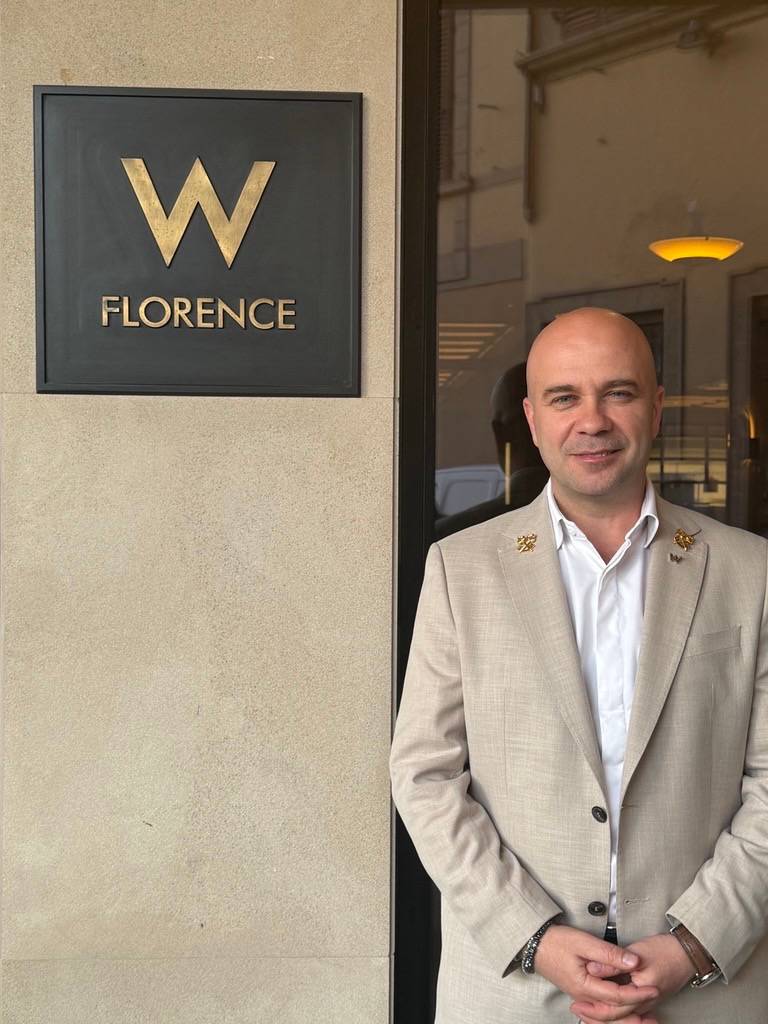 Homem careca de terno bege e camisa branca, com broches dourados, sorri para a câmera em frente à placa preta com W FLORENCE em dourado
