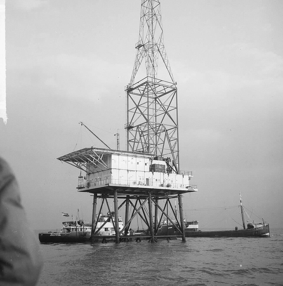 Plataforma de perfuração marítima com torre metálica alta, estrutura principal branca sobre pilares, e dois navios de apoio atracados em águas agitadas sob céu nublado