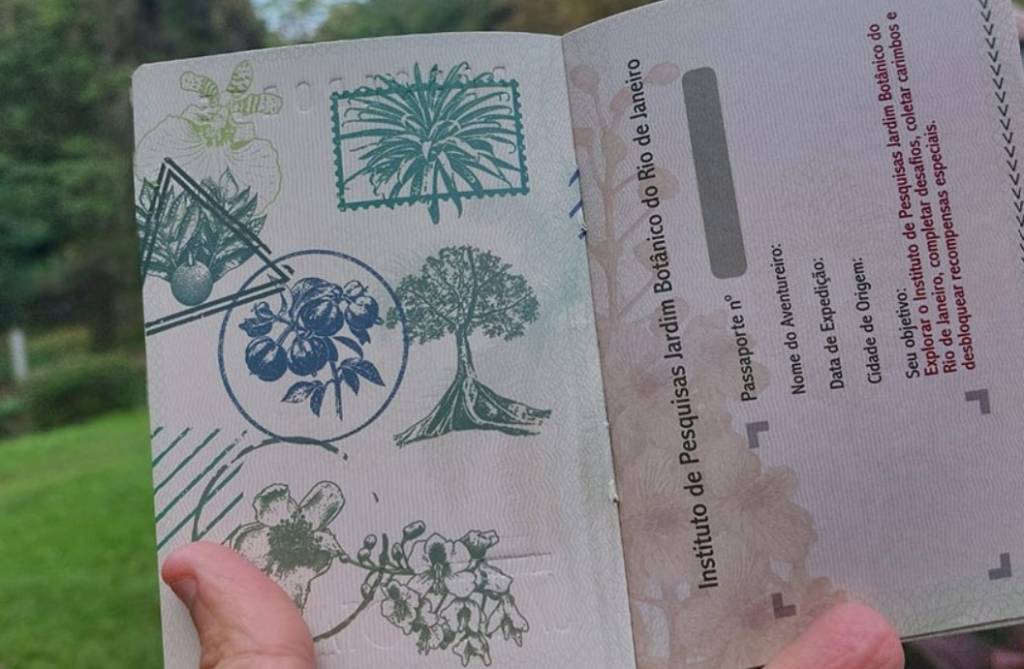 Um passaporte do Instituto de Pesquisas Jardim Botânico do Rio de Janeiro aberto, com carimbos de plantas e flores em tons de verde e azul na página esquerda. A página direita exibe campos para Passaporte nº, Nome do Aventureiro, Data de Expedição, Cidade de Origem e o objetivo de explorar o instituto, coletar carimbos e desbloquear recompensas. Um polegar segura a página inferior esquerda, com fundo verde desfocado