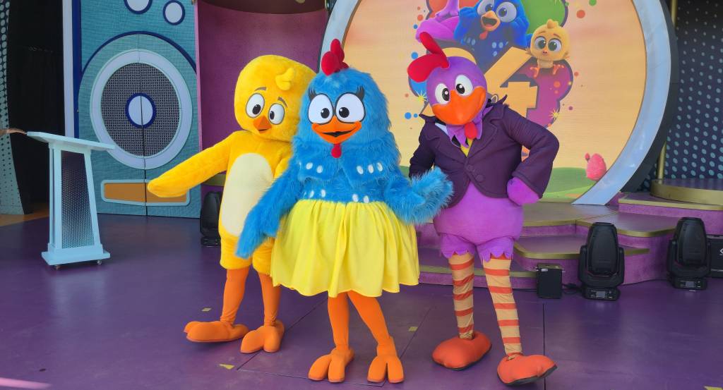 Três personagens da Galinha Pintadinha em fantasias de mascotes no palco: Pintinho Amarelinho, Galinha Pintadinha e o Galo Carijó. Ao fundo, um painel com a imagem de outros personagens e o número 4