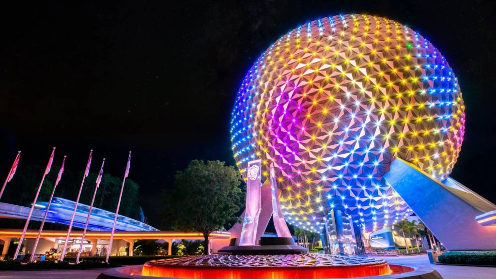 A Spaceship Earth, uma esfera geodésica iluminada em tons de azul, roxo e amarelo, se destaca à noite. À esquerda, bandeiras tremulam e um monorail azul passa rapidamente. No primeiro plano, uma fonte com água avermelhada reflete as luzes. O céu escuro e estrelado completa a cena.