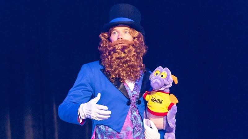 Homem fantasiado de mágico com barba ruiva encaracolada, cartola preta e terno azul, segurando um boneco roxo do Figment, em um palco escuro