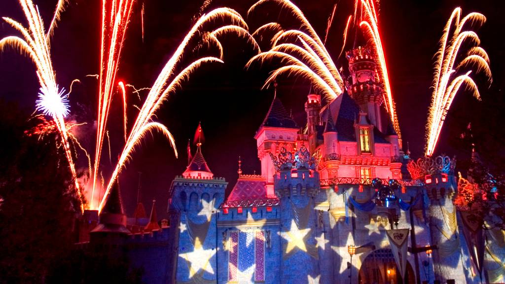 Fogos de artifício vermelhos e dourados explodem sobre um castelo iluminado com projeções de estrelas e bandeiras, criando um espetáculo noturno vibrante
