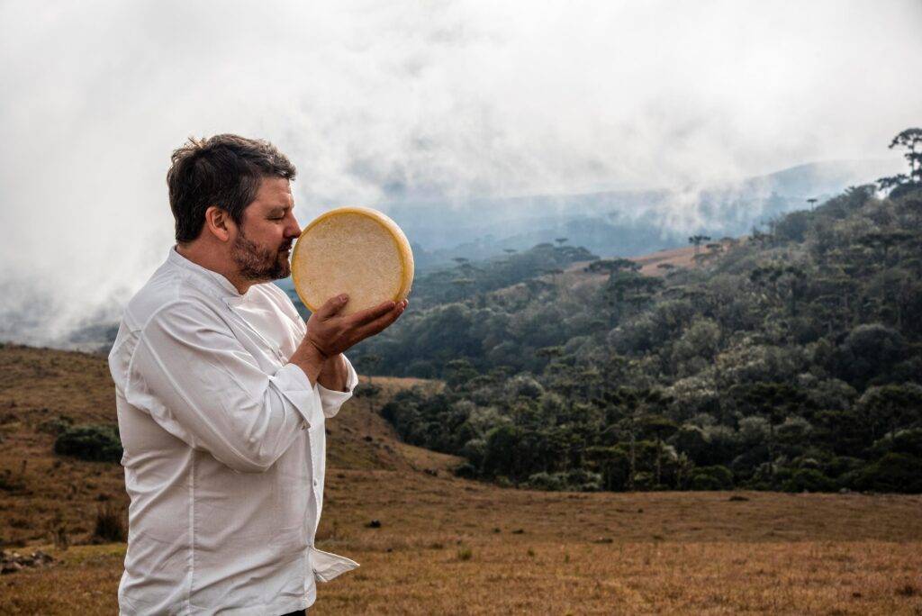 Homem barbudo de camisa branca cheira um queijo redondo e claro, segurando-o com as duas mãos, em um campo com montanhas e árvores ao fundo, sob céu nublado