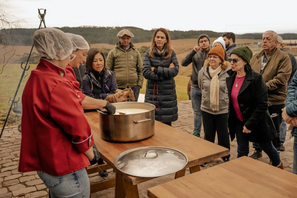 Duas mulheres com toucas de cabelo e aventais vermelhos preparam uma panela grande de comida em uma mesa de madeira, enquanto um grupo de pessoas as observa atentamente em um ambiente externo com colinas ao fundo