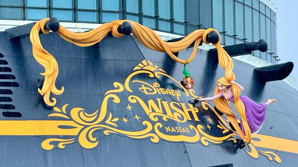 Lateral de um navio de cruzeiro preto com uma faixa amarela e o logotipo dourado Disney Wish Nassau. A princesa Rapunzel, de vestido roxo e cabelos loiros longos, está pendurada no logotipo, com Pascal, seu camaleão verde, e uma paleta de pintura próximos.