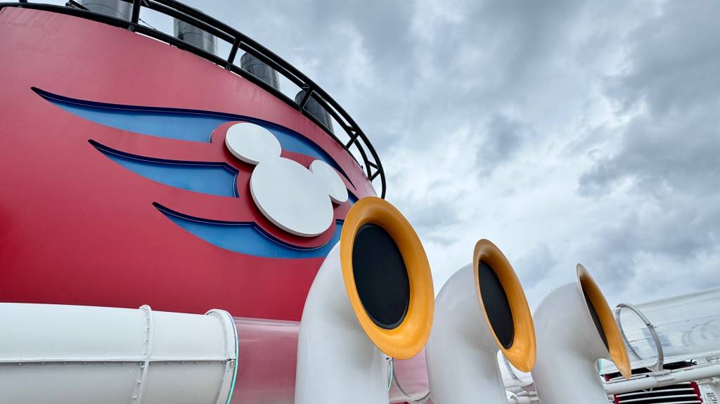 Disney Wish Parte superior de um navio de cruzeiro vermelho e azul com o logo branco do Mickey Mouse, e três toboáguas brancos com bordas amarelas, sob um céu nublado