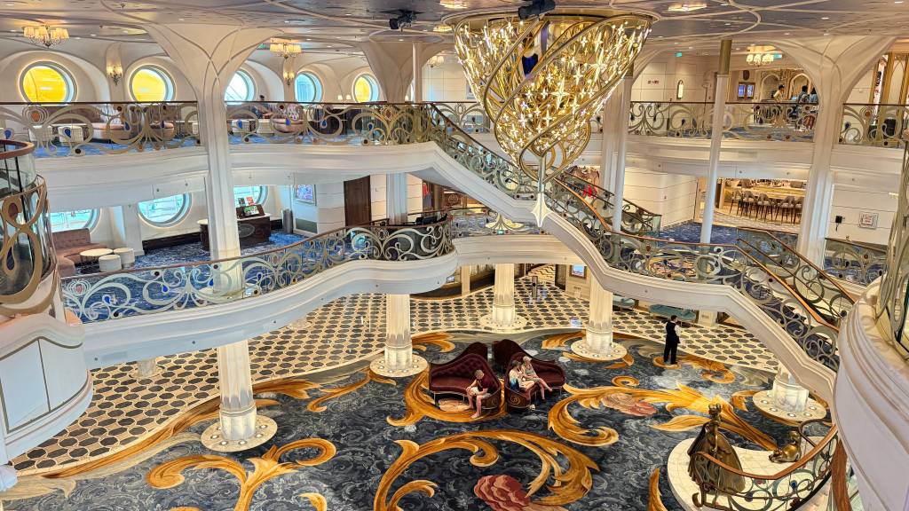 Interior luxuoso de um navio, com escadarias brancas e corrimãos dourados ornamentados. Um lustre dourado imponente pende do teto. No térreo, um tapete azul escuro com detalhes dourados e vermelhos, onde pessoas sentam em sofás curvos de veludo vermelho. Outras pessoas são vistas nos andares superiores, em áreas de convivência com janelas redondas e sofás. Um homem de terno preto está em pé no térreo, próximo a uma estátua dourada. O ambiente é grandioso e elegante