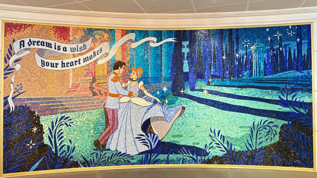 Mosaico colorido de Cinderela e Príncipe Encantado dançando, com a frase A dream is a wish your heart makes em uma faixa branca. O fundo tem um castelo azul e estrelas cintilantes, com folhagens escuras em primeiro plano