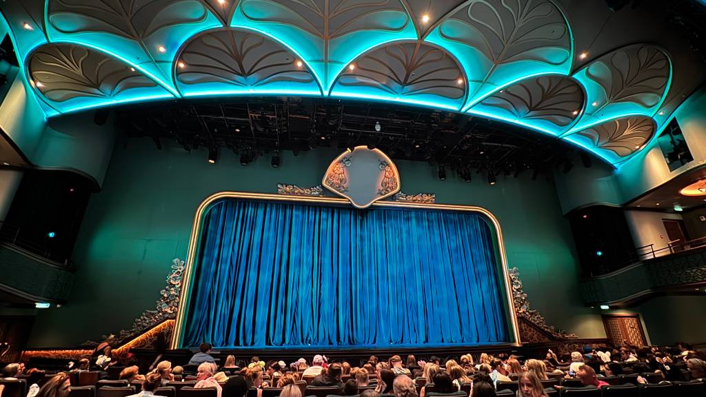 Interior de um teatro com palco fechado por cortinas azuis e uma plateia cheia. O teto tem iluminação azul neon e detalhes dourados em formato de folhas. As paredes são verde-água, com detalhes ornamentados ao redor do palco e um emblema dourado acima das cortinas