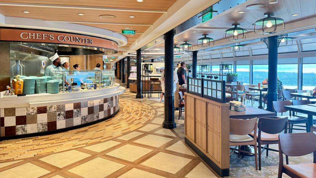 Interior de um restaurante de navio de cruzeiro com balcão de buffet Chefs Counter à esquerda, onde cozinheiros servem. Mesas e cadeiras de madeira preenchem o salão, com grandes janelas ao fundo revelando o mar azul. O teto tem luminárias verdes e o chão é de madeira com padrões geométricos