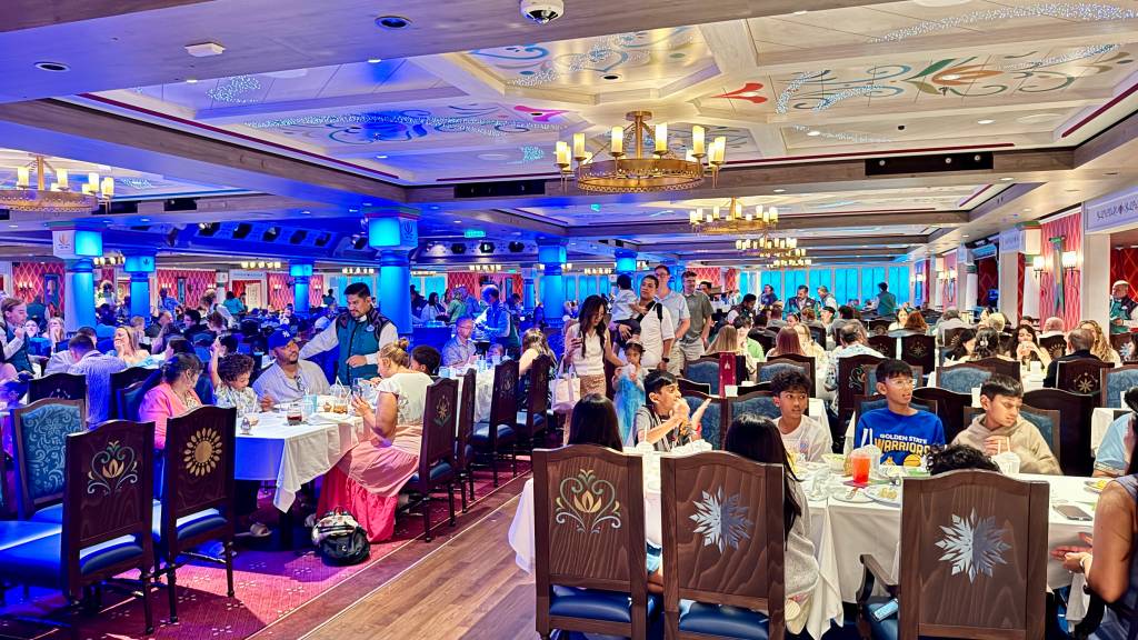 Um restaurante lotado com iluminação azul e dourada, pessoas sentadas em mesas brancas e garçons servindo. O teto tem desenhos em azul e dourado, e lustres pendem sobre as mesas. As cadeiras são de madeira escura com detalhes entalhados.