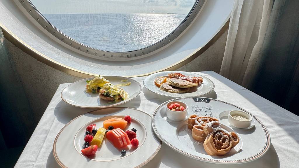 Mesa de café da manhã com ovos Benedict, panquecas com bacon, frutas frescas e waffles em formato de Mickey, com vista para o mar através de uma janela redonda de navio