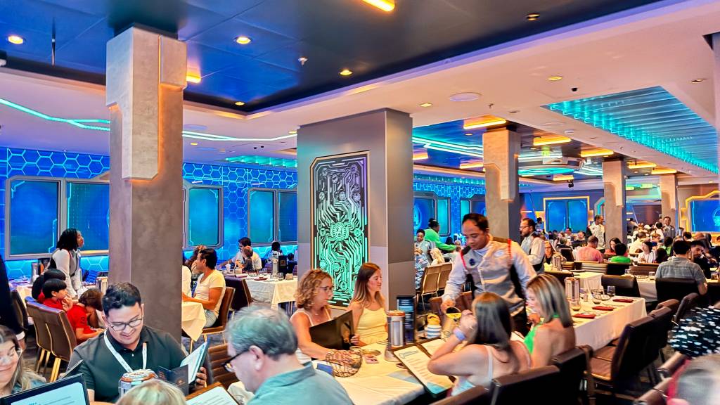 Restaurante futurista com iluminação azul e neon, pessoas sentadas em mesas brancas, garçons servindo, e uma coluna central com arte de circuito eletrônico em tons de verde e roxo