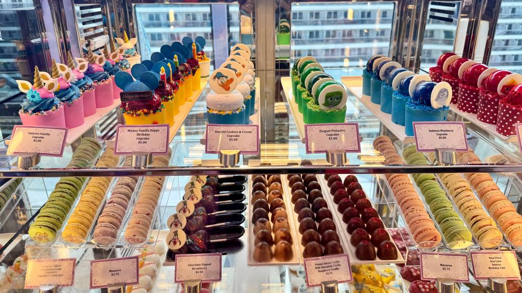 Vitrine de doces com cupcakes temáticos de personagens Disney, como Mickey, Pato Donald, Tristeza e Nojinho, além de macarons coloridos e trufas de chocolate