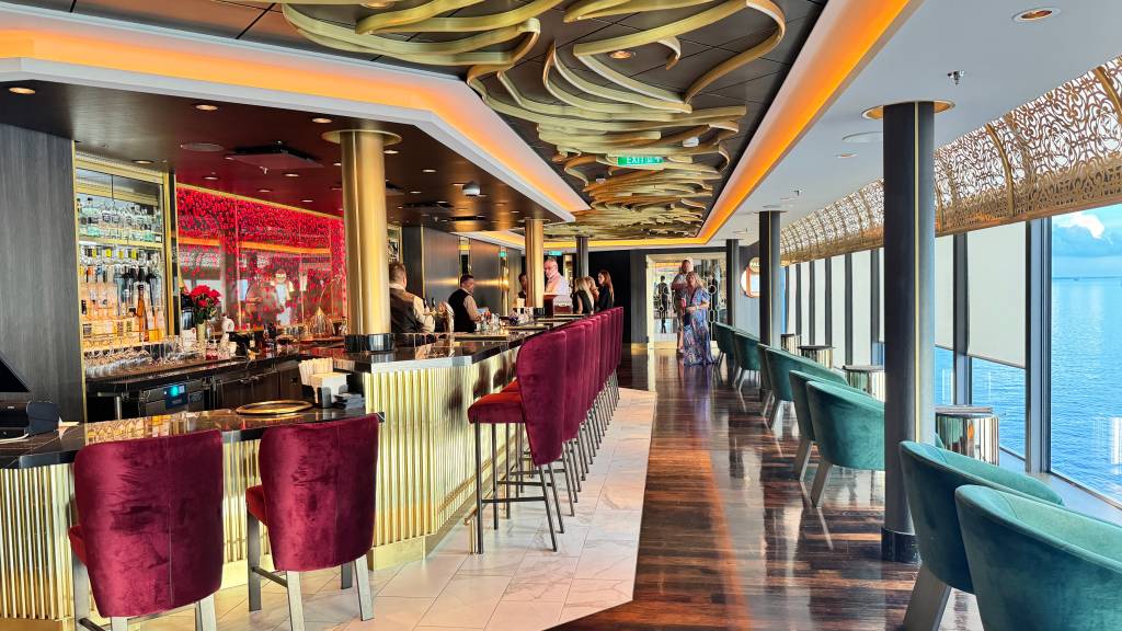 Bar luxuoso em um navio de cruzeiro, com balcão dourado e cadeiras de veludo vinho. À direita, poltronas verde-água e janelas amplas com vista para o mar azul. O teto tem detalhes dourados e iluminação quente. Pessoas conversam no balcão e ao fundo