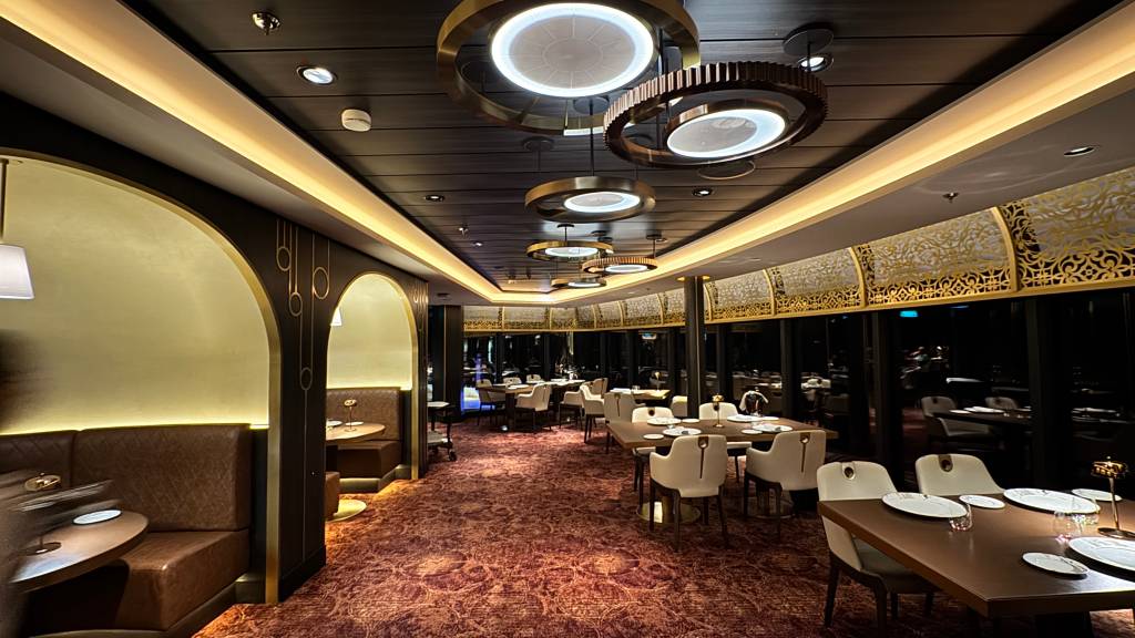 Interior de um restaurante luxuoso com iluminação quente, mesas postas e cadeiras estofadas. O ambiente tem um carpete vermelho, cabines com bancos de couro marrom e arcos dourados nas paredes. O teto escuro possui luminárias circulares modernas e detalhes em dourado nas janelas.