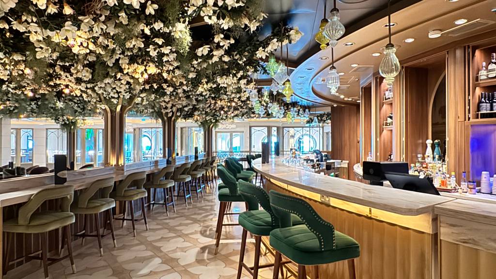 Interior de um bar luxuoso com balcão curvo de mármore claro e banquetas estofadas em veludo verde-oliva. O teto é decorado com folhagens e flores brancas, simulando árvores, e lustres pendentes. Ao fundo, janelas arqueadas e prateleiras de madeira com bebidas.