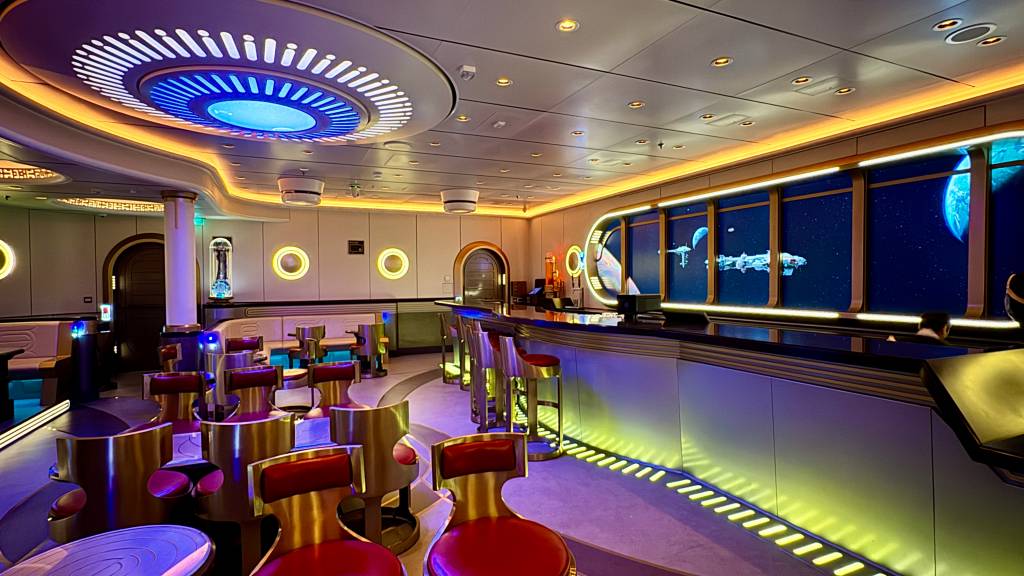 Interior de um bar futurista com iluminação neon azul e amarela. Há um balcão longo com bancos vermelhos e dourados, e mesas redondas com cadeiras semelhantes. Ao fundo, janelas virtuais exibem uma nave espacial e planetas no espaço sideral. O teto tem um grande círculo iluminado em azul e branco, e as paredes são claras com detalhes circulares iluminados