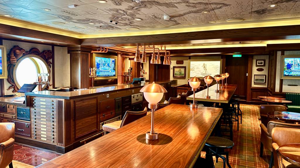 Interior de um bar com decoração náutica, mesas e cadeiras de madeira escura, luminárias de cobre e teto com mapa antigo. Um balcão de bar com torneiras de chope e uma janela redonda com moldura de polvo entalhada. Duas TVs exibem notícias.