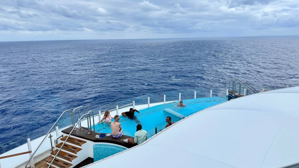 Pessoas em uma piscina de borda infinita em um navio, com o vasto oceano azul e céu nublado ao fundo. Quatro pessoas estão na água, e duas sentadas na borda, desfrutando do passeio.