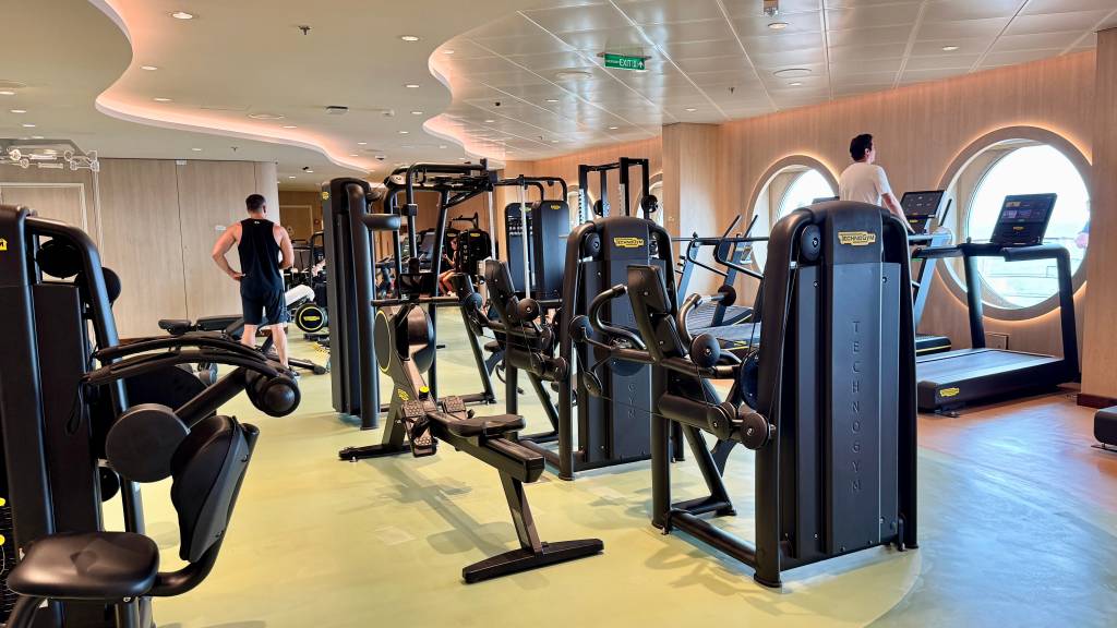Academia moderna com equipamentos Technogym, incluindo esteiras e máquinas de musculação. Dois homens se exercitam, um de camiseta branca na esteira e outro de regata preta no centro. O ambiente tem paredes claras, janelas redondas e iluminação embutida no teto curvo