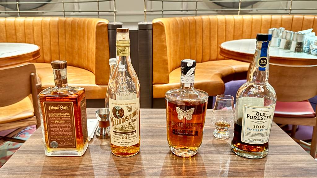 Quatro garrafas de uísque de diferentes marcas e formatos, com um copo de degustação e um medidor de dose, sobre uma mesa de madeira em um bar com estofados de couro marrom ao fundo