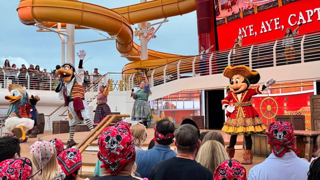 Personagens Disney, Pateta e Pato Donald, vestidos de piratas, interagem com o público em um palco de navio de cruzeiro. Minnie Mouse pirata acena à direita, enquanto pessoas assistem, algumas com bandanas do Mickey. Um toboágua amarelo e uma tela vermelha com AYE, AYE, CAPT ao fundo completam a cena festiva