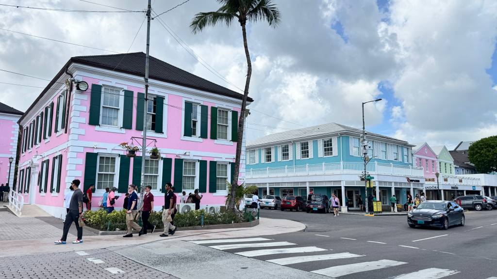 Pessoas caminham por uma rua com edifícios coloridos em tons pastel, como rosa e azul, com janelas de venezianas verdes. Uma palmeira alta se destaca no centro, e carros estão estacionados ou em movimento na rua, sob um céu nublado com nuvens esparsas