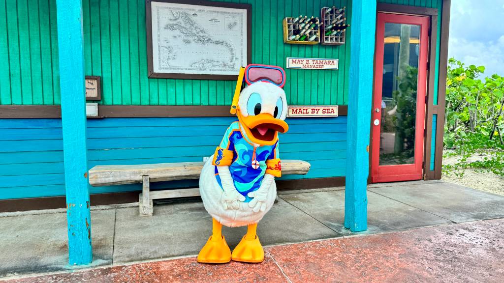 Pato Donald fantasiado de mergulhador, com máscara e snorkel na cabeça, vestindo uma camisa de mergulho azul e laranja, em frente a uma construção de madeira azul e verde com um mapa na parede e uma porta vermelha, ao lado de um banco de madeira