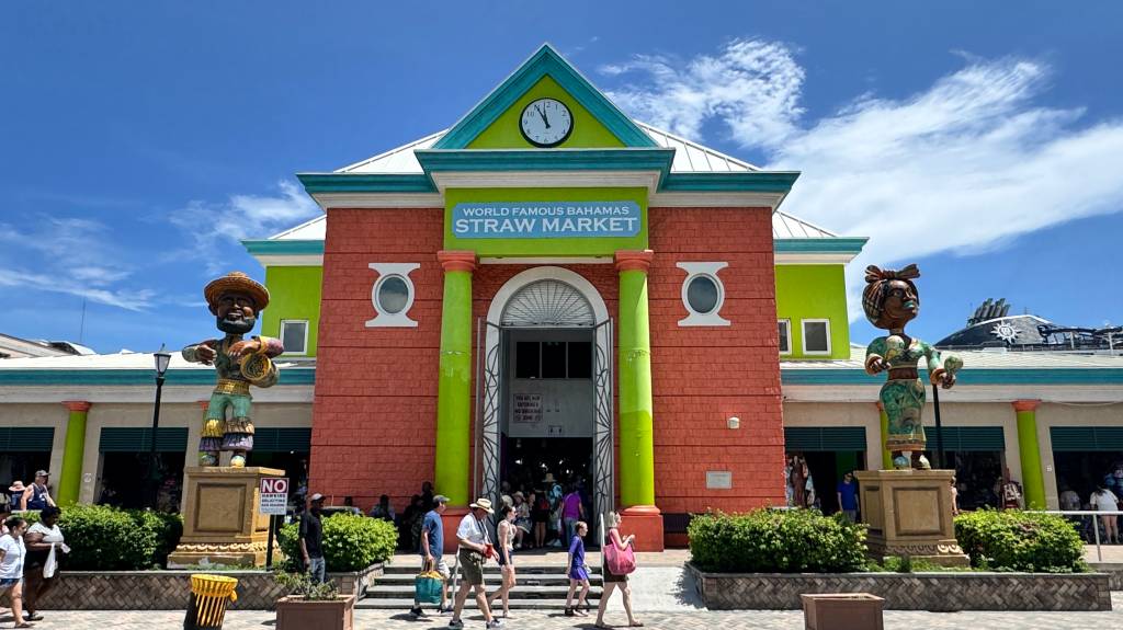 Fachada colorida do World Famous Bahamas Straw Market, com paredes de tijolos vermelhos, detalhes em verde-limão e azul-turquesa, e um relógio no topo. Duas estátuas de figuras caribenhas flanqueiam a entrada, enquanto pessoas caminham em frente ao mercado sob um céu azul com nuvens esparsas