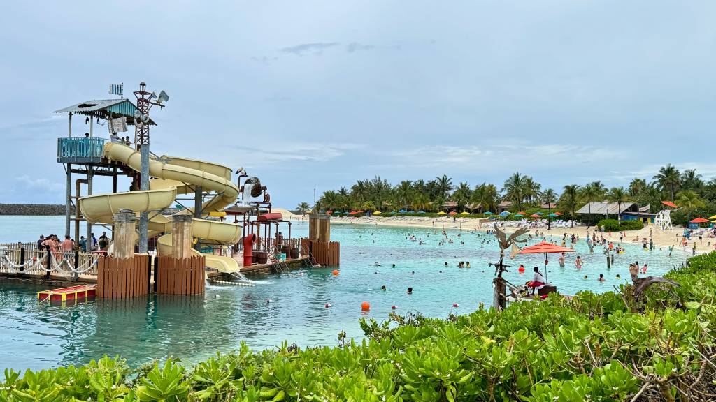 Parque aquático com toboáguas amarelos em uma lagoa de água azul-turquesa, com muitas pessoas nadando e relaxando na praia de areia branca, cercada por coqueiros e guarda-sóis coloridos sob um céu nublado