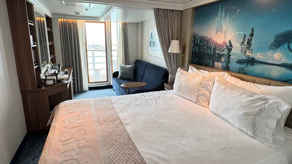 Quarto de navio de cruzeiro com cama de casal, sofá azul, varanda e uma pintura de castelo mágico na parede. A cama tem lençóis brancos e uma manta com a palavra Wish em relevo. À esquerda, uma mesa com espelho e garrafas.