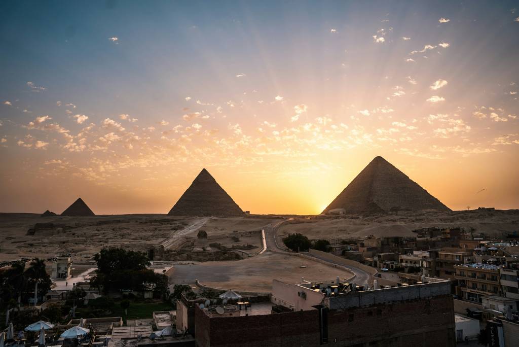 Três pirâmides egípcias no deserto, com o sol se pondo entre as duas maiores, iluminando o céu em tons de laranja e azul. Nuvens esparsas e raios de sol atravessam o céu. Em primeiro plano, telhados de edifícios e uma estrada sinuosa.