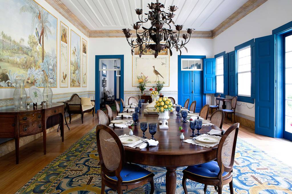 Sala de jantar luxuosa com mesa de madeira escura posta para doze pessoas, cadeiras com encosto de palha e assento azul. Paredes brancas com murais de paisagens tropicais e pássaros, portas e janelas com venezianas azuis. Um lustre de ferro forjado escuro pende do teto branco. Um tapete azul e dourado cobre o chão de madeira
