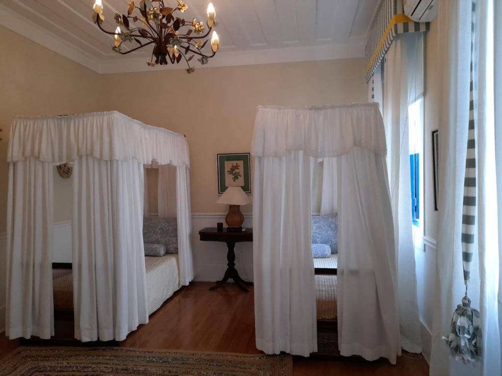 Quarto com duas camas de solteiro com dossel de cortinas brancas, piso de madeira e tapete estampado. Um lustre floral de ferro forjado pende do teto branco, e uma mesa de cabeceira escura com abajur está entre as camas. Uma janela com cortinas listradas e persiana amarela aparece à direita