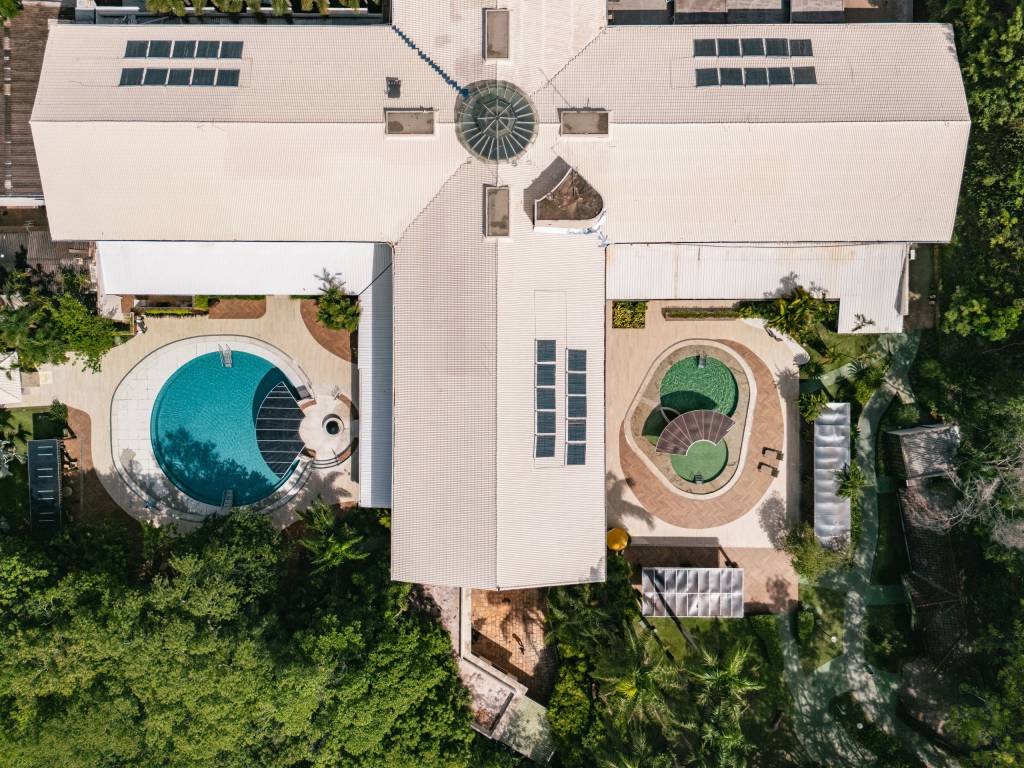 Vista aérea de um complexo de lazer com telhados claros, duas piscinas e muita vegetação. À esquerda, uma piscina azul em formato de C com deck de madeira. À direita, duas piscinas menores de formato oval com água esverdeada. Painéis solares e claraboias no telhado complementam a estrutura.
