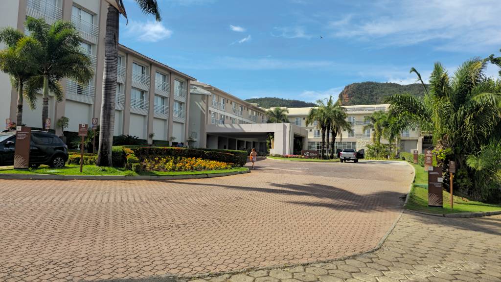 Entrada de um hotel com fachada bege e sacadas brancas, palmeiras altas e canteiros floridos. Uma pessoa caminha na via de paralelepípedos, e carros estão estacionados. Ao fundo, montanhas verdes sob céu azul com nuvens esparsas