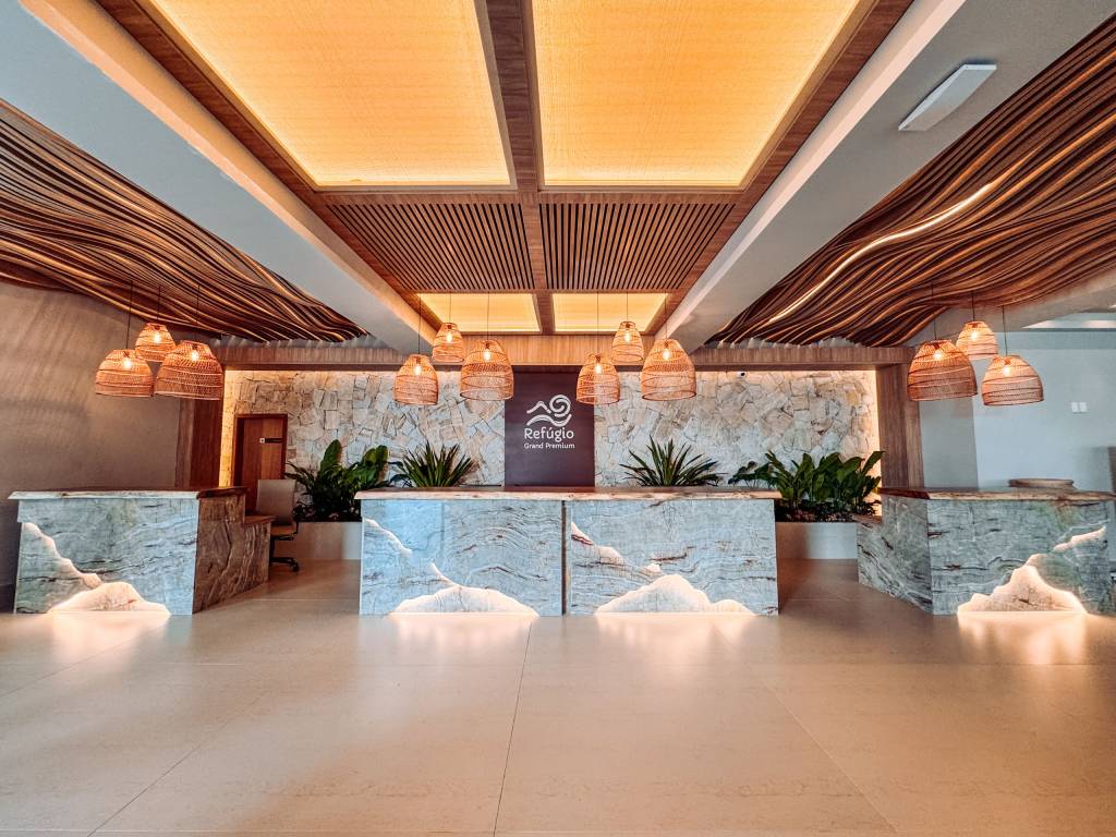 Recepção de hotel com balcões de mármore iluminados, teto de madeira ondulada e luminárias de palha penduradas. Ao fundo, uma parede de pedra com plantas verdes e o logo Refúgio Grand Premium