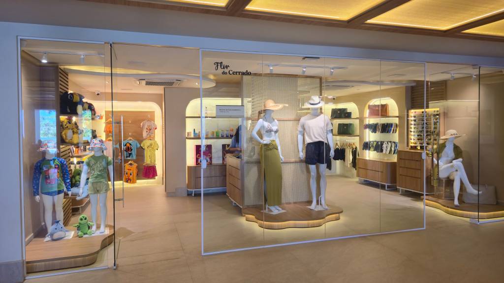 Vitrine da loja Flor do Cerrado com roupas de praia e acessórios. À esquerda, manequins infantis com camisetas coloridas e chapéus. No centro, manequins adultos exibem biquíni, saia longa, camiseta e shorts. À direita, um manequim sentado com chapéu e óculos.