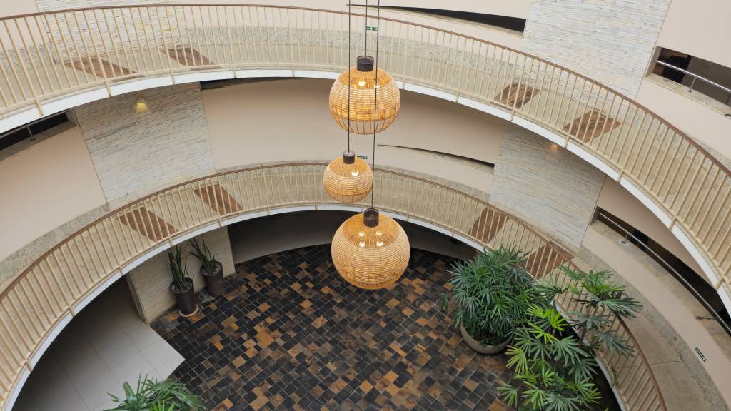 Vista aérea de um saguão circular com três andares, corrimãos bege e paredes claras. Três grandes luminárias esféricas de vime pendem do teto, iluminando o piso de ladrilhos pretos e marrons. Plantas verdes em vasos decoram o ambiente