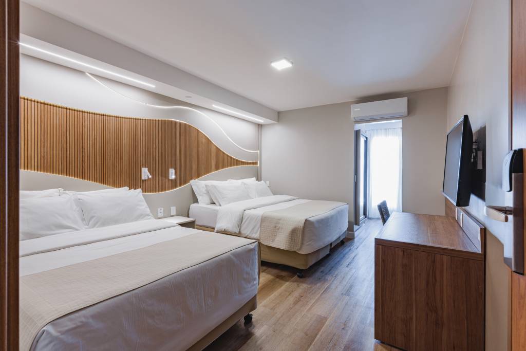 Quarto de hotel moderno com duas camas de casal, lençóis brancos e colchas bege. A cabeceira tem painéis de madeira verticais e iluminação embutida. À direita, uma TV preta montada na parede acima de uma escrivaninha de madeira escura. Uma porta-janela aberta revela uma cortina branca e luz natural. O piso é de madeira clara