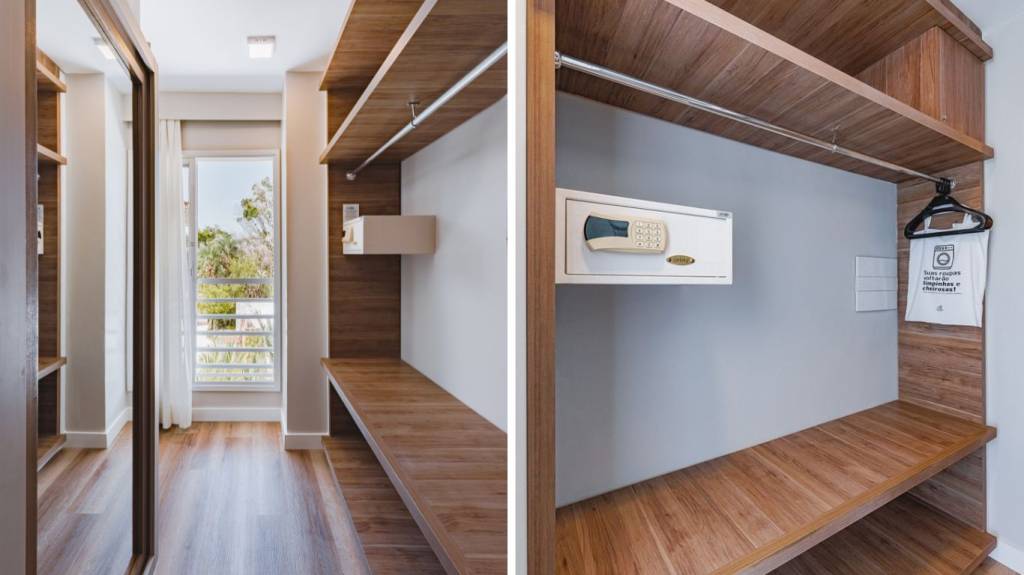 Um closet moderno com piso e prateleiras de madeira clara, paredes brancas e um espelho de corpo inteiro. Há um cofre digital branco embutido e um saco de lavanderia pendurado em um cabide. Ao fundo, uma janela com vista para árvores e um céu azul