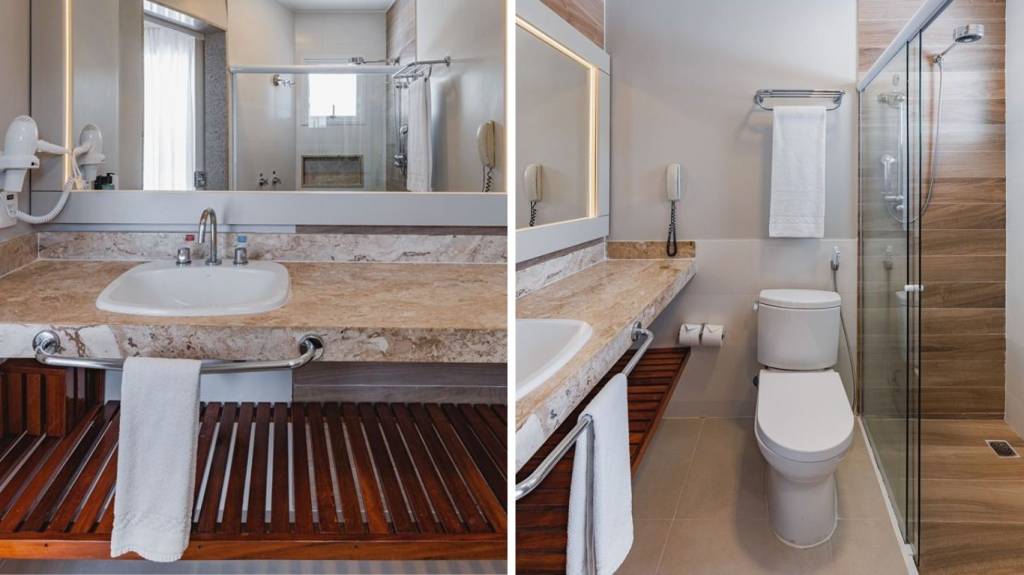 Banheiro moderno com bancada de mármore bege, pia branca, espelho grande e secador de cabelo. À direita, vaso sanitário branco e box de vidro com chuveiro, parede revestida em madeira. Toalhas brancas penduradas em barras metálicas.