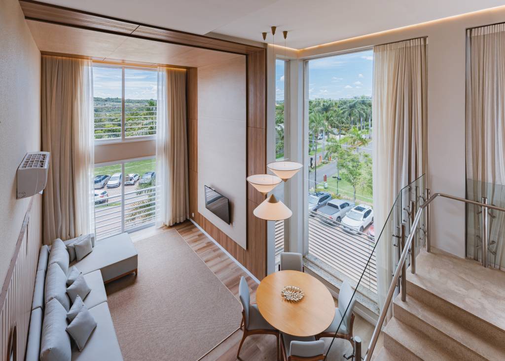 Sala de estar e jantar em um apartamento duplex, com sofá cinza em L, tapete bege, TV na parede de madeira, mesa de jantar redonda com quatro cadeiras e luminárias pendentes. Grandes janelas com cortinas claras revelam um estacionamento e uma rua arborizada com palmeiras. Uma escada com corrimão de vidro e metal aparece à direita