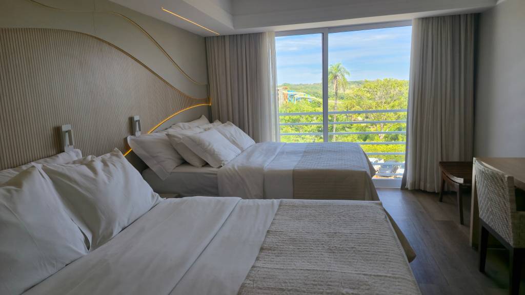Quarto de hotel moderno com duas camas de solteiro arrumadas, lençóis brancos e colchas bege. A cabeceira tem detalhes ondulados e iluminação indireta. Uma grande janela com cortinas claras revela uma paisagem verde com árvores e céu azul. À direita, uma mesa de madeira com cadeira de palha e um banco de madeira escura. O piso é de madeira clara.