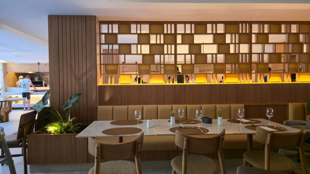 Interior de restaurante moderno com mesas de mármore, cadeiras de madeira e estofamento mostarda. Ao fundo, um chef de camisa 10 prepara alimentos em cozinha aberta, e uma parede decorativa de madeira e espelhos reflete o ambiente