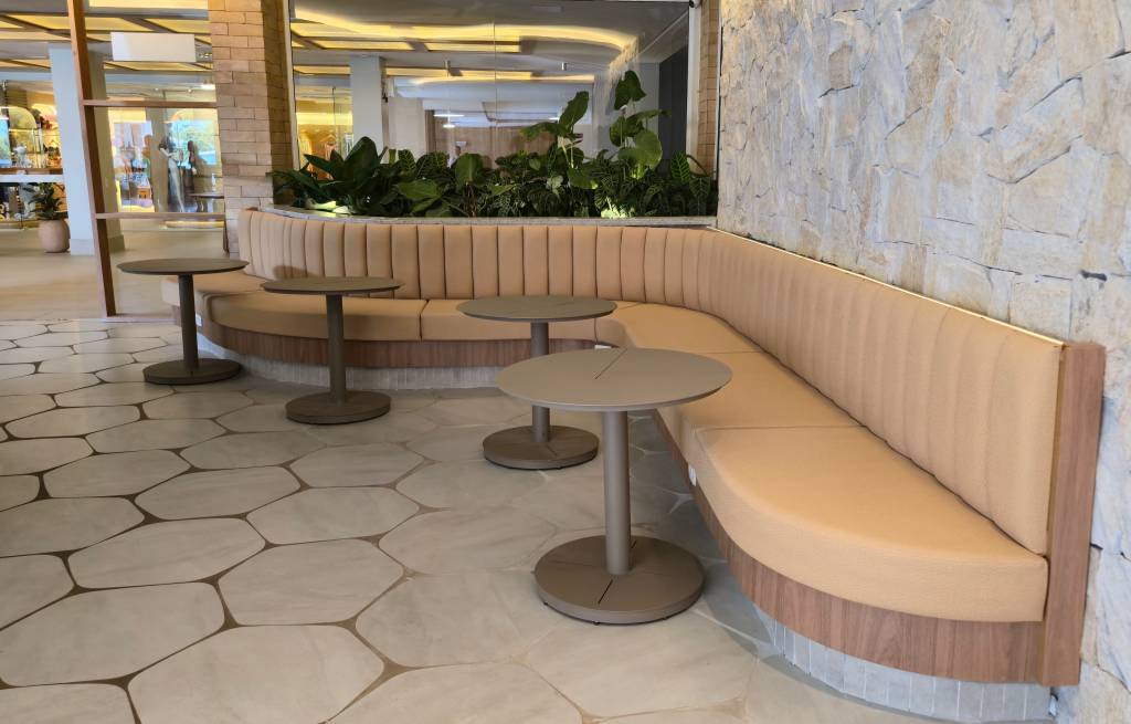 Área de espera moderna com um longo sofá curvo bege e mesas redondas cinza, em frente a uma parede de pedras claras e um piso de hexágonos. Plantas verdes decoram o fundo