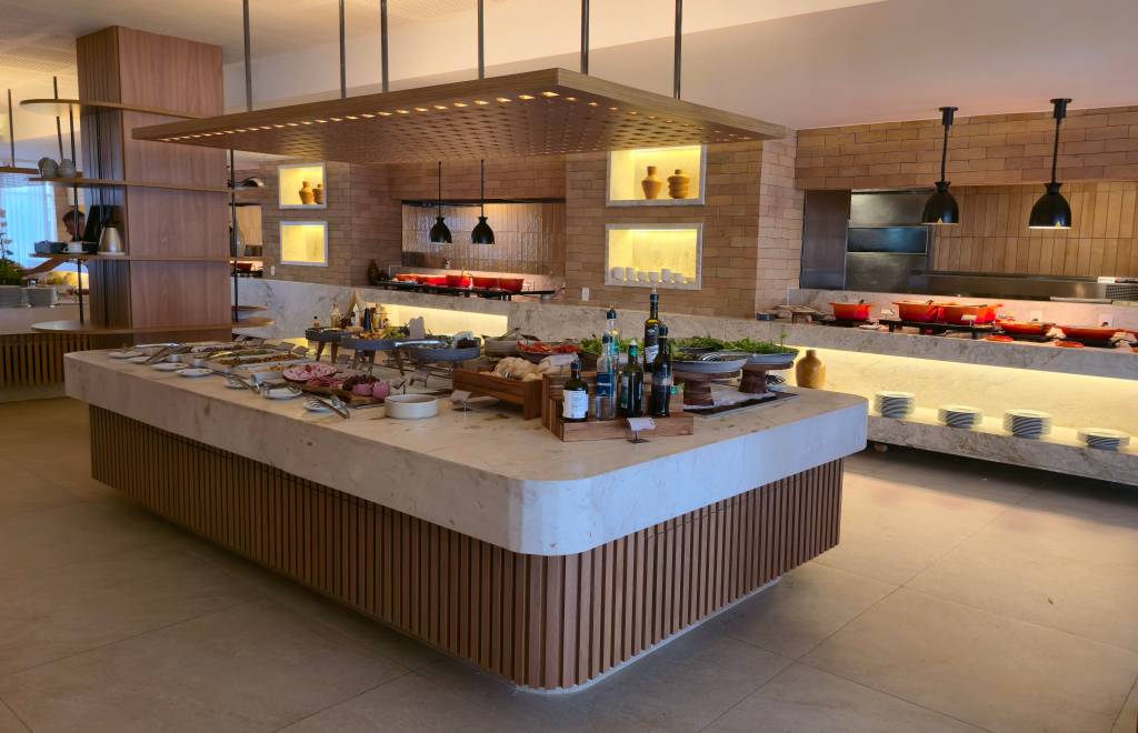 Um buffet de restaurante moderno com uma ilha central de mármore e madeira, exibindo saladas, frios e azeites. Ao fundo, panelas vermelhas em um balcão iluminado, com paredes de tijolinho e luminárias pendentes pretas